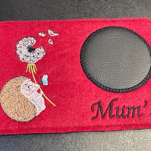 May include: Red felt coaster with a black leather circle in the center, embroidered with a hedgehog, a dandelion, and a butterfly. The text "Mum's" is embroidered below the circle.