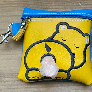 May include: A yellow zippered pouch with a blue trim and a key chain. The pouch has an embroidered design of a bear's rear end with a white plastic bag inside.