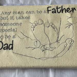 May include: Canvas art piece with embroidered text and a hand-drawn illustration. The text reads "Any man can be a Father but it takes someone special to be a Dad." The illustration depicts a pair of baby feet in a hand.