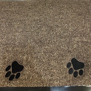 May include: A rectangular doormat with a textured, speckled brown surface. Two black paw prints are near the bottom edge. The mat is designed to trap dirt and debris from shoes.