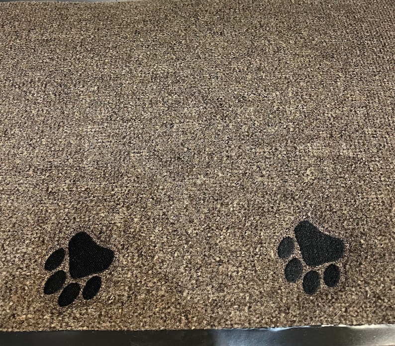 May include: A rectangular doormat with a textured, speckled brown surface. Two black paw prints are near the bottom edge. The mat is designed to trap dirt and debris from shoes.