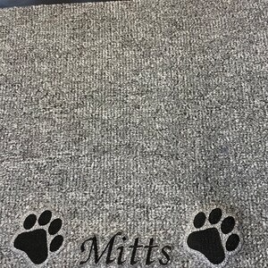 May include: Gray doormat with a black border. The mat features two black paw prints and the name "Mitts" embroidered in a script font. The mat has a textured surface.