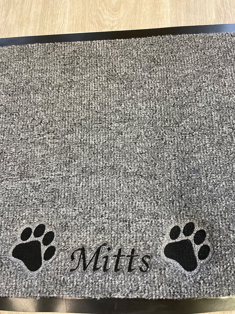 May include: Gray doormat with a black border. The mat features two black paw prints and the name "Mitts" embroidered in a script font. The mat has a textured surface.