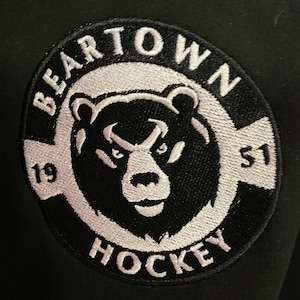 Könnte beinhalten: Schwarzer und weißer gestickter Aufnäher mit einem Bärenkopf in der Mitte. Der Text "Beartown Hockey" befindet sich um den Bärenkopf herum. Die Zahlen "19" und "51" befinden sich auf beiden Seiten des Bärenkopfes.