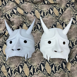 Könnte beinhalten: Zwei weiße, traditionelle japanische Oni-Masken mit Hörnern und übertriebenen Gesichtszügen. Die Masken sind aus glattem, weißem Material gefertigt und haben eine leicht strukturierte Oberfläche.