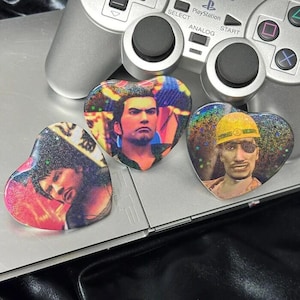 Puede incluir: Tres pegatinas holográficas en forma de corazón con imágenes de personajes, colocadas sobre un mando de PlayStation plateado. Las pegatinas tienen un efecto brillante y probablemente son coleccionables o decorativas.
