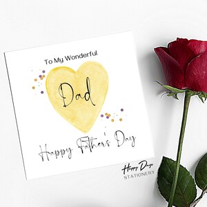 May include: A white card with a yellow heart that says "Dad" inside. The heart is surrounded by gold and purple confetti. The card says "To My Wonderful Dad Happy Father's Day" and is signed "Happy Days Stationery".