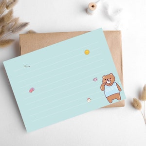 Può includere: Un blocco note a righe blu chiaro con un'illustrazione di un orso bruno che si spazzola i denti. L'orso indossa una camicia a righe blu e bianche. Ci sono altre piccole illustrazioni di cartoni animati sul blocco note, tra cui un sole, una tazza e un fiore.