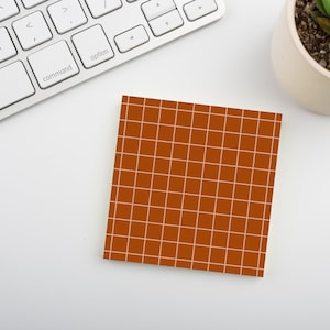 Op de afbeelding: Een vierkant, oranje plaknotitieblok met een wit rasterpatroon. Het notitieblok ligt op een wit oppervlak naast een wit toetsenbord en een kleine vetplant in een pot.