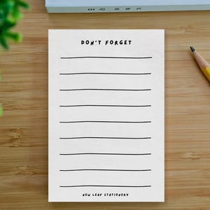 Op de afbeelding: Een witte notitieblok met de woorden "DON'T FORGET" bovenaan en "NEW LEAF STATIONERY" onderaan. Het notitieblok heeft horizontale lijnen om op te schrijven. Een gele potlood ligt op een houten oppervlak.