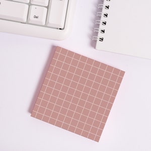 Op de afbeelding: Een roze geblokte plakbriefje op een wit oppervlak. Het briefje is leeg en klaar om op te schrijven.