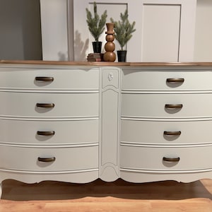 May include: White dresser with six drawers and a wood top. The dresser has ornate details and brass handles.