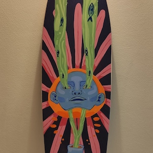 Könnte beinhalten: Ein Longboard-Skateboard mit einem farbenfrohen Design. Das Design zeigt ein blaues Gesicht mit grünen Ranken, die aus den Augen und dem Mund wachsen. Die Ranken sind von rosa Blütenblättern und grünen Blättern mit kleinen blauen Fischen umgeben. Der Hintergrund ist dunkelblau.