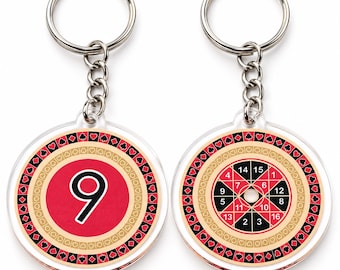 May include: Two circular keychains with silver metal rings and chains. One keychain features a large white number 9 on a red background. The other displays a numbered roulette-style wheel with red and black segments. Both have decorative borders.