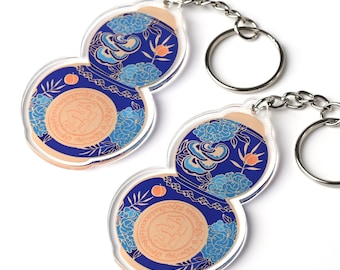 May include: Two gourd-shaped keychains with intricate designs. The keychains feature a blue background with light blue floral patterns and peach-colored accents. Each keychain has a silver-colored chain and key ring.