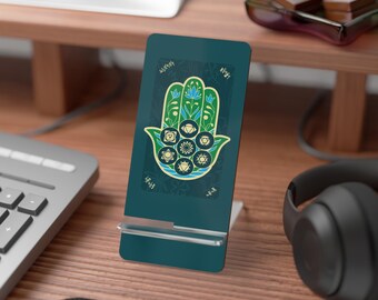 May include: A teal phone stand featuring a Hamsa hand design with seven chakra symbols. The hand is green and blue with floral accents. The stand is on a wooden desk with a keyboard and headphones.