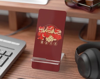 May include: A burgundy phone stand with a gold and red illustration of three mythical creatures. The stand is on a wooden desk with a keyboard, headphones, and other items. The illustration has Chinese characters.