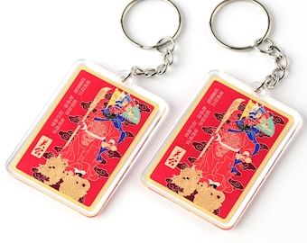May include: Two red and gold keychains with a Chinese character and a picture of a person in traditional clothing. The keychains are attached to silver key rings.