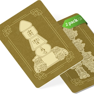 May include: Two gold-colored cards with intricate designs. One card features a stacked, stylized shape with script, while the other has a decorative border and the text "2 pack" on a green banner. The cards have a detailed, patterned background.