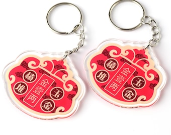 May include: Two red and gold Chinese New Year keychains with the words "金壹两" and "堆" on them. The keychains are made of clear acrylic and have a silver chain.
