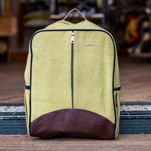 May include: A green canvas backpack with a brown leather base and a black zip. The backpack has a small black tag with the text "LIVING JUTE" on it.