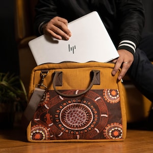 May include: Mustard-coloured laptop bag with a brown strap and handles. The bag has a decorative panel with a circular pattern in shades of brown, orange, and white. A silver laptop is partially visible inside the bag.