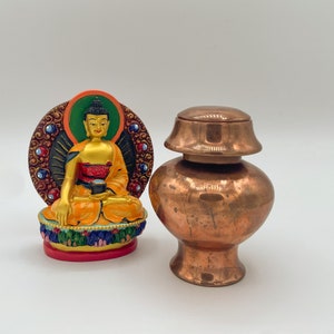 Op de afbeelding: Een goud-oranje Boeddha-beeld met een decoratieve, kleurrijke achtergrond. Daarnaast een koperkleurige urn met deksel. Het beeld en de urn staan op een wit oppervlak.