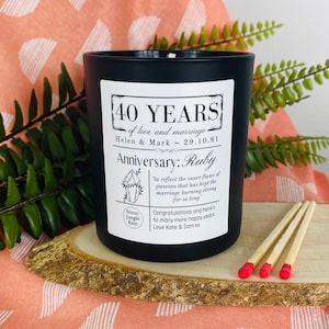 May include: Black candle with a white label that reads "40 Years of love and marriage Helen & Mark ~ 29.10.81 Anniversary: Ruby" with a small graphic of a ruby crystal and the text "Scent: Jungle Rain" in a circle. Three red-tipped matches are on a wooden surface.