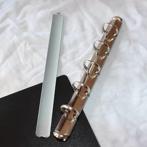 Pode incluir: Um mecanismo de encadernação prateado com cinco argolas e uma espinha dorsal de metal prateado. O mecanismo está sobre uma superfície preta.