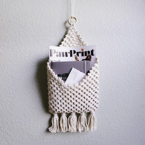 May include: Cream-colored macrame wall organizer with a triangular top, a wooden ring, and a fringe at the bottom. The organizer holds a magazine with the text "PawPrint", a gray notebook, and a white envelope.