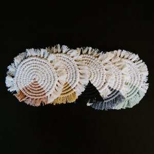 May include: Five round crocheted coasters with fringe edges. The coasters are in shades of beige, light brown, black, gray, and green.