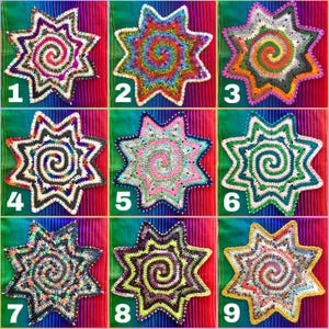 May include: Nine colorful, star-shaped crochet doilies with spiral centers are displayed. Each doily features a unique color palette, including vibrant rainbow hues, greens, pinks, and purples. The doilies are arranged on a striped, multi-colored fabric.