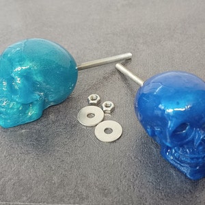 May include: Two blue skull-shaped cabinet knobs with silver screws and washers. The skull on the left is a teal blue and the skull on the right is a darker blue.