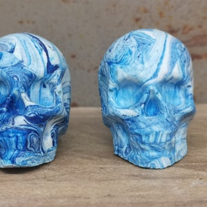 May include: Two blue and white marbled skull sculptures. The skulls are made of a smooth, white material and are painted with a swirling blue and white pattern.