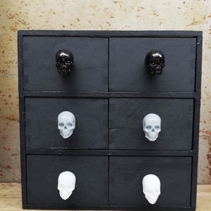 Peut inclure: Une commode en bois noir avec six tiroirs. Chaque tiroir a une poignée en forme de crâne. Les poignées sont noires, grises et blanches.