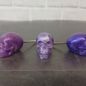 May include: Three purple skull-shaped bottle stoppers with a metallic sheen. The skulls are different shades of purple, with one being a lighter shade with a white swirl.