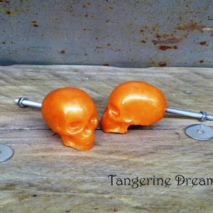 May include: Two orange skull-shaped cabinet knobs with silver metal hardware. The knobs are attached to a wooden surface.