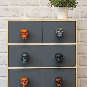 May include: A wooden chest of drawers with six drawers, each featuring a skull-shaped knob. The chest is painted a dark grey and has a light wood trim. The knobs are in various colours, including black, orange, and brown.
