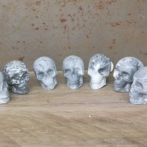 May include: Seven small, white and black marbled skull figurines. The skulls are arranged in a row on a wooden surface.