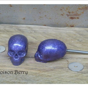 Peut inclure: Deux poignées de tiroir en forme de crâne en paillettes violettes avec des vis et des rondelles argentées. Les poignées de crâne sont sur une surface en bois.