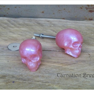 May include: Two pink skull-shaped cabinet knobs with a pearlescent finish. The knobs are attached to a metal rod with a washer and nut. The text "Carnation Breeze" is visible on the wooden surface.
