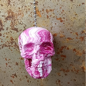 May include: A skull-shaped pull chain pendant with a swirling pink and white pattern. The pendant hangs from a silver ball chain against a textured, rusty metal background.
