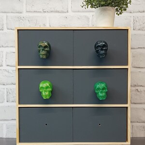 May include: A wooden dresser with four drawers, each with a skull-shaped knob. The top two drawers have black skull knobs, and the bottom two drawers have green skull knobs. The dresser is sitting on a white brick wall background.