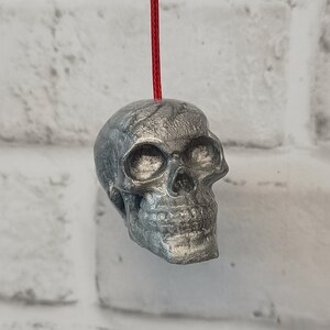 May include: A silver skull pendant with a red string attached. The skull has a detailed design with a textured surface.