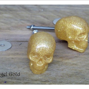Peut inclure: Deux poignées de tiroir en forme de crâne recouvertes de paillettes dorées avec une vis argentée. Les poignées en forme de crâne sont sur une surface en bois. Angel Gold.