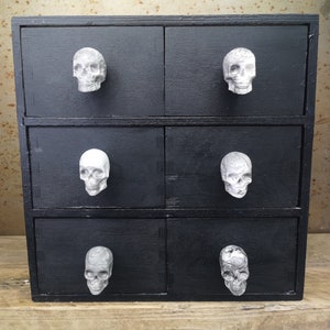 May include: A black wooden chest of drawers with six drawers. Each drawer has a white skull-shaped knob.