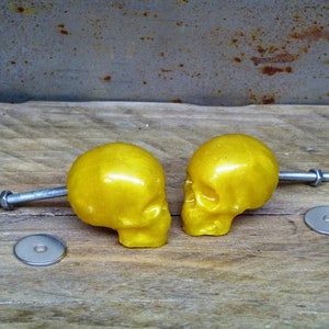 May include: Two golden skull-shaped cabinet knobs with silver hardware. Each skull is mounted on a bolt with a washer and nut. The knobs are displayed on a weathered wooden surface.