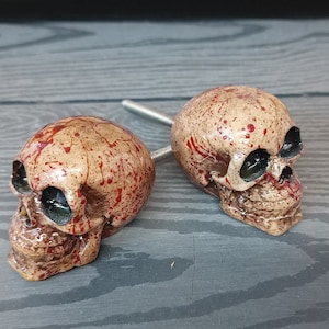 May include: Two small, detailed, ceramic skull drawer pulls with a beige base and red splatter paint.