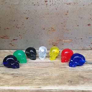 May include: Seven colourful skull-shaped drawer pulls in various colours including green, black, white, yellow, red, and blue. The pulls are arranged in a semi-circle on a wooden surface.