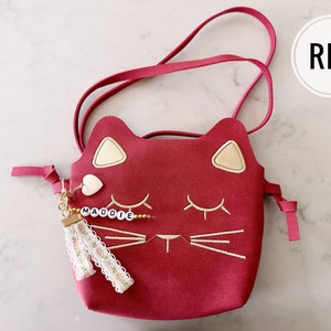 May include: Red cat-shaped crossbody purse with a gold chain and a tassel charm. The charm has a small heart and the name "MADDOIE" spelled out in white beads.